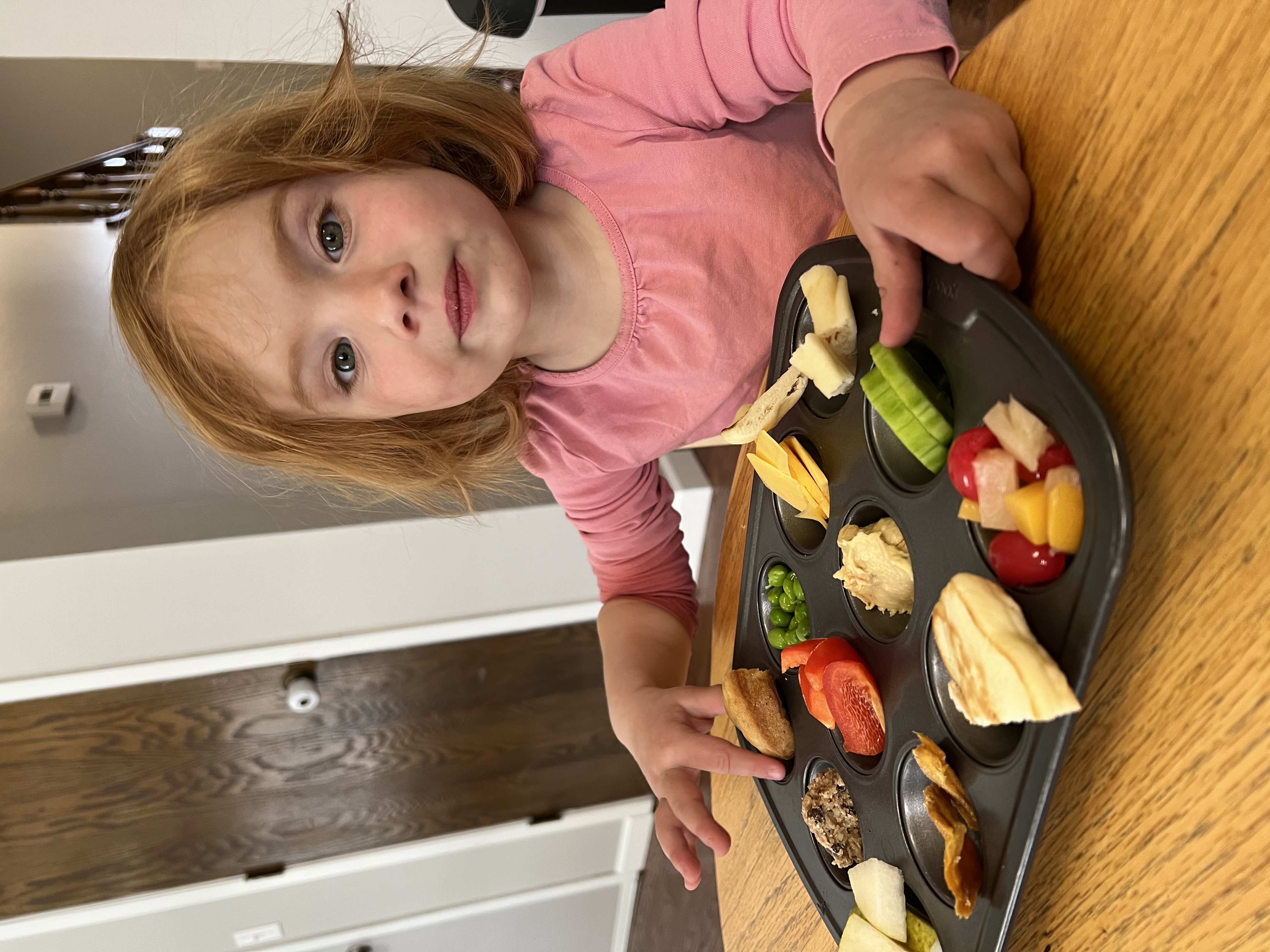 Toddler girl smiling with a muffin pan meal on the table